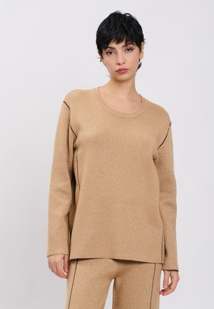 Kvinde med kort sort hår iført en beige striktrøje og matchende bukser med mørke sømdetaljer, stående mod en ensartet lys baggrund.