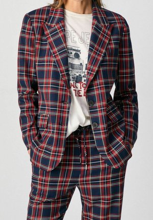 Kvinde iført navyblå og rødternet blazer og matchende bukser med hvid grafisk T-shirt stoppet ind, hænderne i lommerne.