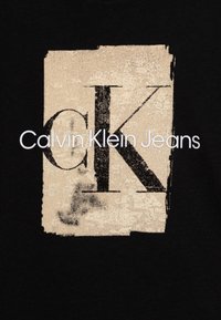 Černé bavlněné tričko s velkým béžovým obdélníkem a opotřebovaným černým logem "CK" umístěným nad názvem značky "Calvin Klein Jeans" v bílém písmu.