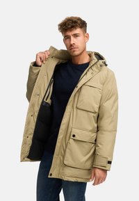 Beige isolert parka-jakke med hette, har to store frontlommer, knappelukking, og en myk indre fôr. Stylet med en blå genser under.