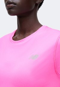 Κοντινό πλάνο ενός ατόμου που φοράει φωτεινό ροζ αθλητικό μπλουζάκι New Balance με στρογγυλή λαιμόκοψη και αντανακλαστικό λογότυπο στο στήθος.