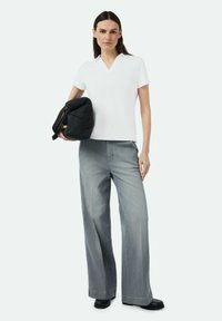 Weiße V-Ausschnitt-Bluse kombiniert mit weit geschnittenen, hellgrauen Jeans. Das Model hält eine schwarze, gesteppte Tasche. Schwarze Schuhe und minimale Accessoires.