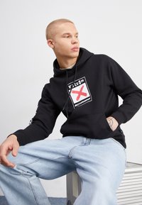 Sudadera negra con un parche de logo en el frente, que presenta una 'X' roja sobre el logo. Combinada con unos vaqueros anchos de color azul claro, el tejido parece suave y casual.