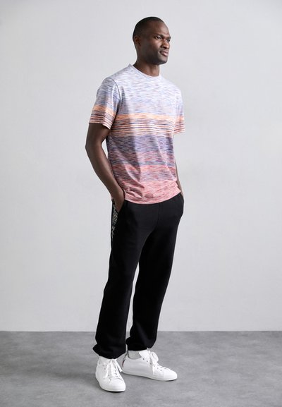 T-shirt multicolore à manches courtes avec des motifs horizontaux, associé à un pantalon de survêtement noir et des baskets blanches, se tenant devant un arrière-plan neutre.