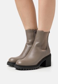 Bottines en cuir gris avec une finition texturée, panneaux élastiques latéraux et une semelle extérieure noir épaisse. Talon empilé pour plus de hauteur et de stabilité.