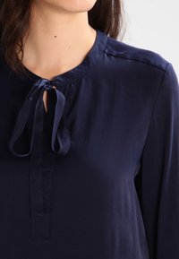 Kvinna i en marinblå blus med knytkrage, visad från halsen till överkroppen mot en enfärgad bakgrund.