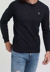 Pull tricoté bleu marine avec un motif texturé, des poignets et un ourlet côtelés, un col rond, et un petit accent logo. Associé à un jean bleu clair.