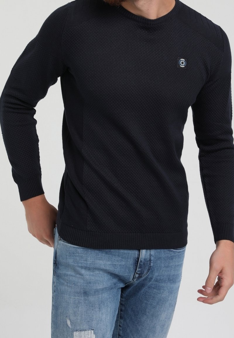 Pull tricoté bleu marine avec un motif texturé, des poignets et un ourlet côtelés, un col rond, et un petit accent logo. Associé à un jean bleu clair.