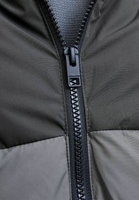 Dunkle, voluminöse Jacke mit einem kontrastierenden hellgrauen Einsatz, versehen mit einem robusten schwarzen Reißverschluss und einer glatten Stofftextur.