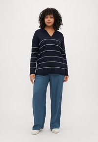 Marineblauwe pullover met witte horizontale strepen, V-hals, lange mouwen, gecombineerd met lichtblauwe wijde broek en beige instap schoenen.