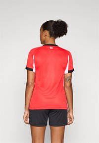 Rød sportstrøye med svarte detaljer, korte ermer og rund hals. Båret med svarte shorts som viser en jevn tekstur og atletisk passform.