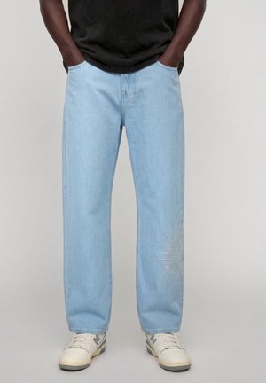 Persoon draagt lichtblauwe straight-leg jeans met zon-borduursel bij de enkel, witte sneakers en een zwart shirt, handen in de zakken.