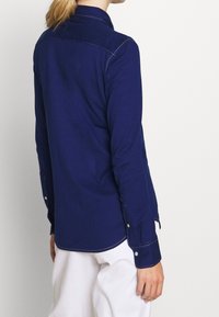 Person wearing a long-sleeve navy blue shirt with white stitching and white pants, shown from the back against a plain background.