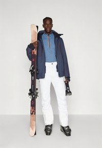 Síruha, amely sötétkék dzsekit, kék felsőt narancssárga cipzárral, fehér nadrágot és fekete síbakancsokat tartalmaz, miközben egy sílécet fog, amelyen kötés van.