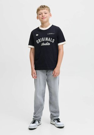 T-shirt noire avec des accents blancs, arborant le texte "ORIGINALS studio". Associée à un jean gris clair et des baskets blanches avec des lacets noirs.