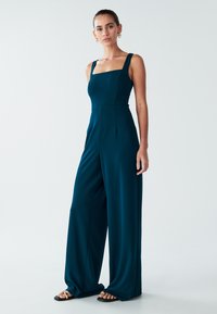 Turkizni kombinezon s kvadratnim izrezom, debelimi naramnicami in širokimi hlačnicami. Izdelan iz gladkega blaga z rahlim prepogibom in čistimi šivi.