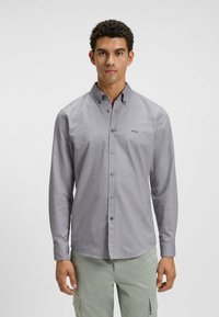 Camisa de manga larga gris con botones y cuello, que presenta una suave textura y un pequeño logotipo en el área izquierda del pecho. Botones plateados.