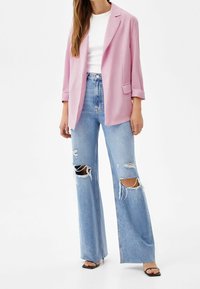 Light pink blazer, fitted design, open front. Worn over a white top. Distressed wide-leg blue jeans with several large rips. Black sandals.