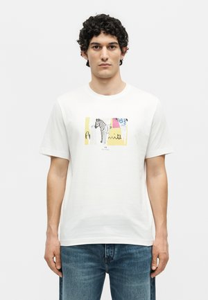 Άνδρας που φοράει λευκό κοντομάνικο T-shirt με πολύχρωμο γραφικό ζέβρας και κείμενο μπροστά, συνδυασμένο με μπλε τζιν, σε απλό φόντο.