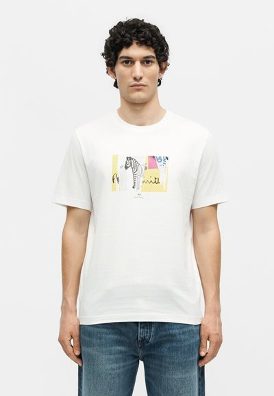 Άνδρας που φοράει λευκό κοντομάνικο T-shirt με πολύχρωμο γραφικό ζέβρας και κείμενο μπροστά, συνδυασμένο με μπλε τζιν, σε απλό φόντο.