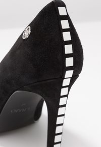 Zapato de tacón alto de ante negro con un logo metálico, que presenta un patrón a cuadros blanco a lo largo del tacón y un diseño de punta afilada.