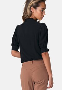 Młoda kobieta w czarnej bluzce z puff-sleeve i beżowych spodniach, zwrócona w bok z odwróconą głową, na jednolitym jasnym tle.