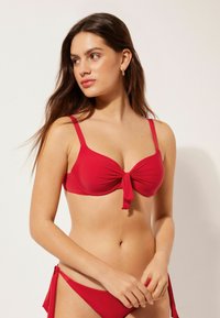 Bikini rouge avec un détail noué au centre. Fabriqué en tissu lisse et extensible. Fines bretelles et bas taille basse avec liens sur les côtés.