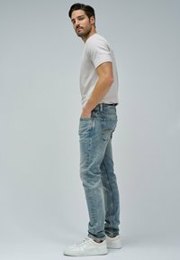 Camiseta gris clara, jeans azules desgastados y zapatillas blancas. Los jeans tienen un ligero desgaste. El modelo posa de lado contra un fondo simple.