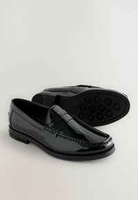 Svarta lackläderloafers med en slät yta, dekorativt band över framsidan, samt en flexibel gummisula med cirkulära mönster.