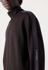 Fekete, cipzáros dzseki magas gallérral, texturált oldalsó panelek és hímzett "Calvin Klein" logó a bal ujjon. Simább anyag.