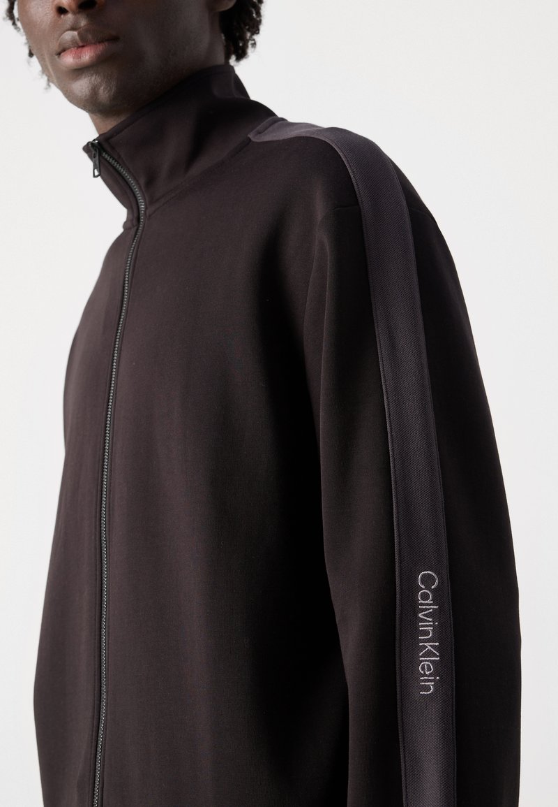 Giacca nera con zip e colletto alto, pannello laterale testurizzato e logo ricamato "Calvin Klein" sulla manica sinistra. Tessuto liscio.