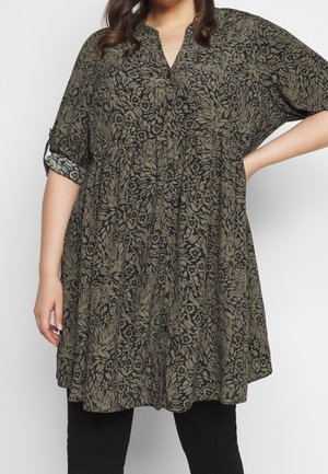 Kobieta nosząca luźną sukienkę z przodu na guziki, z podwiniętymi rękawami i oliwkowozielonym kwiatowym wzorem liści na czarnym tle.