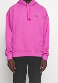 Sweat à capuche rose avec une poche kangourou à l'avant. Présente un petit logo Levi's noir sur la poitrine et des poignets côtelés aux manches.