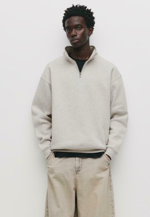 Light gray zip-up sweatshirt with a high collar, ribbed cuffs, and a relaxed fit, worn over a black shirt, paired with beige trousers.