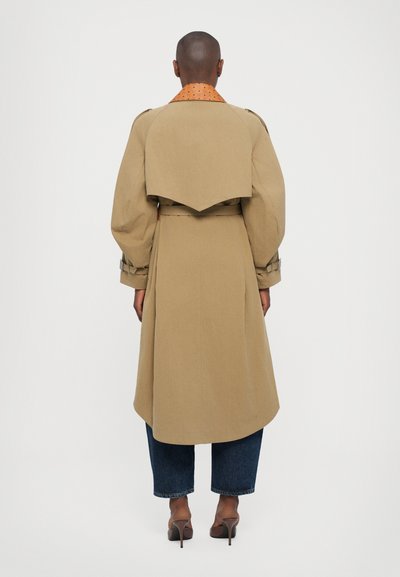 Bež trench coat vööga, millel on üle mõõtme varrukad ja kumer tagune detail. Sisemine krae on oranž ja muster.