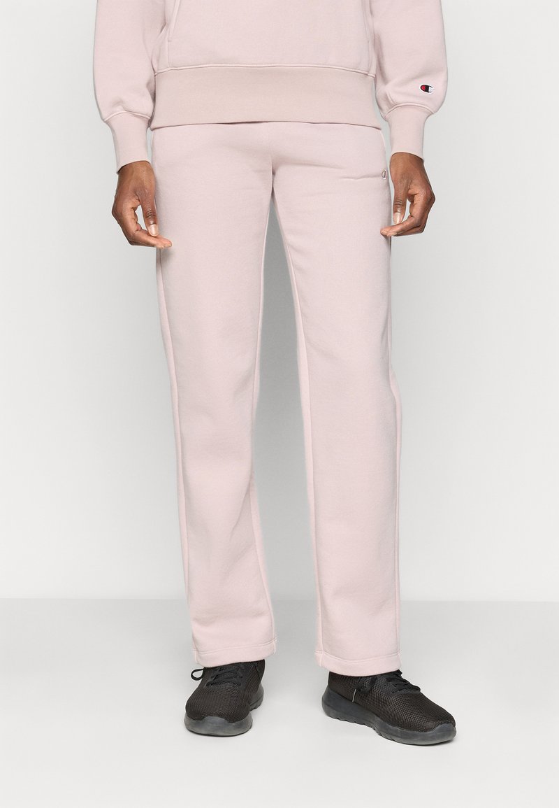Modèle masculin portant un pantalon de survêtement rose clair et un sweat-shirt assorti, debout avec des baskets noires contre un fond clair uni.