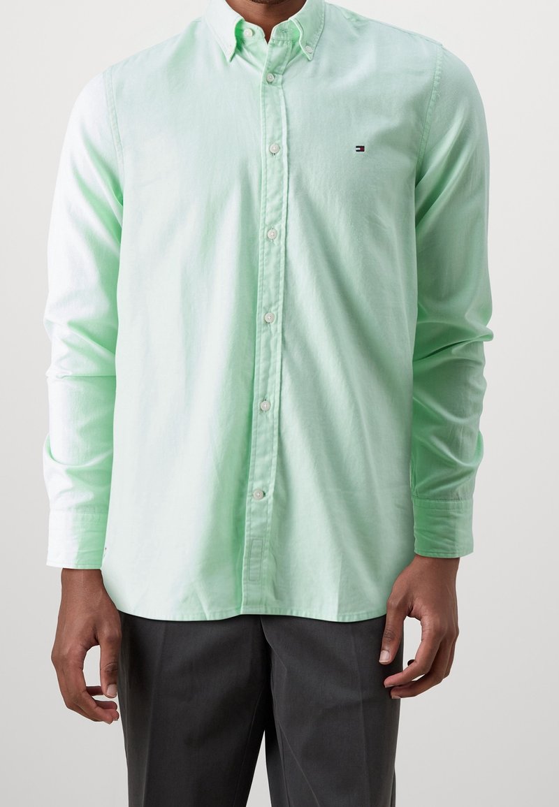 Camisa de botones verde claro de algodón, con cuello, mangas largas y un pequeño logo en el lado izquierdo del pecho, combinada con pantalones grises.