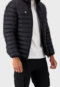 Giacca a vento nera con design trapuntato orizzontale, cappuccio, chiusura con zip e patch logo esagonale sul lato sinistro del petto. Indossata sopra una camicia bianca.