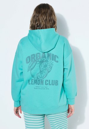 Osoba nosząca turkusową bluzę z kapturem z napisem „Organic Lemon Club” i grafiką cytryny na plecach, w połączeniu z turkusowo-białymi wzorzystymi spodniami.