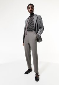 Grijze blazer met een slim fit, gecombineerd met een gestreepte coltrui en grijze high-waisted broek. Zwarte schoenen met unieke ontwerpelementen.