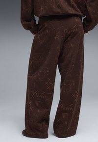 Personne portant un pantalon marron ample et un haut à manches longues assorti avec des motifs abstraits et des logos Puma, debout devant un fond gris.