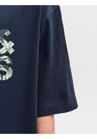 Κοντινό πλάνο μανικιού κοντομά νικης μπλούζας σε ναυτικό μπλε χρώμα, με μερικά πράσινα και μαύρα φύλλα σε μοτίβο στο στήθος.