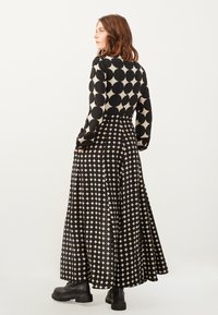 Lang jurkje met een zwart en crème geometrisch patroon, met een aansluitende bodice, lange mouwen en een vloeiende rok. Effen zwarte schoenen.