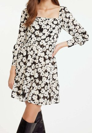Femme portant une robe noire à imprimé floral blanc, col carré, manches longues et bottes noires montantes jusqu'aux genoux, posée avec une main sur la hanche.
