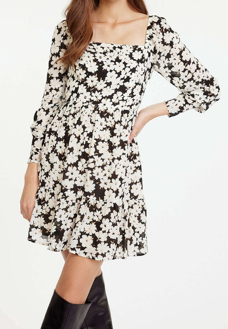 Femme portant une robe noire à imprimé floral blanc, col carré, manches longues et bottes noires montantes jusqu'aux genoux, posée avec une main sur la hanche.