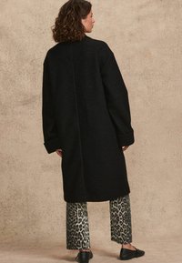 Langer schwarzer Wollmantel mit lässigem Schnitt, breiten Ärmeln und einer Mittelnaht, kombiniert mit weitem Hosen im Leopardenmuster.