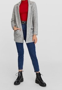Grauer Strickblazer mit Schalkragen, schwarze Jeans und schwarze Schnürstiefel. Verfügt über Fronttaschen und eine taillierte Silhouette. Roter Rollkragenpullover darunter.