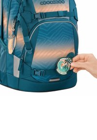 Türkise Rucksack mit geometrischem Muster, ausgestattet mit einem gewebten Patch mit der Aufschrift "OCEAN VIBES" und einem Palmbaum-Design. Mehrere Riemen und Reißverschlüsse sind sichtbar.