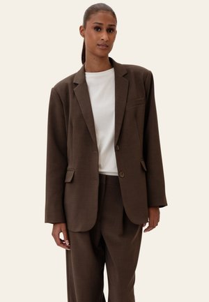 Kobieta o ciemnej karnacji i włosach związanych do tyłu ubrana w oversize'ową brązową marynarkę i dopasowane spodnie, na białej koszuli, stoi na jednolitym tle.