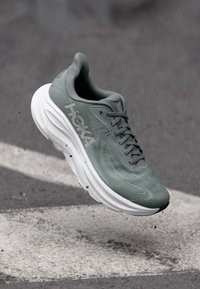 Γκρι αθλητικό παπούτσι Hoka με λευκή σόλα, αιωρούμενο πάνω από ανάγλυφο άσφαλτο με λευκές βαμμένες γραμμές.